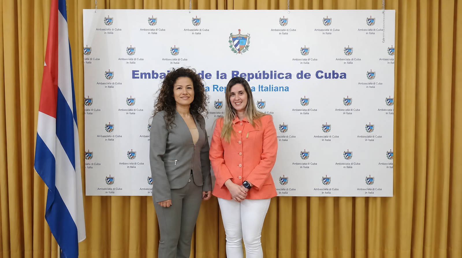 Due donne, una in completo grigio e una con giacca arancione, davanti al fondale dell’Ambasciata di Cuba.